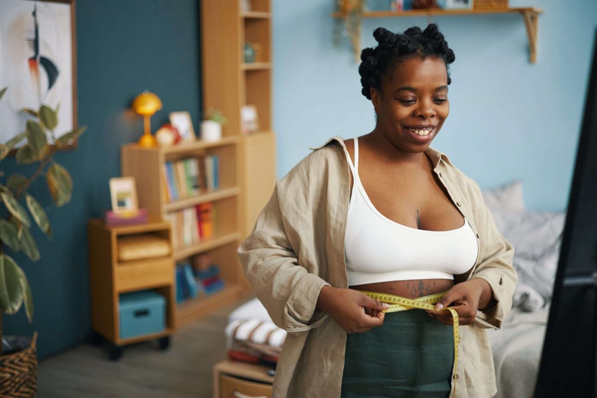 Smiling Black person measuring waistline with measuring tape while standing in stylish and brightly-lit living room setting filled with modern decor